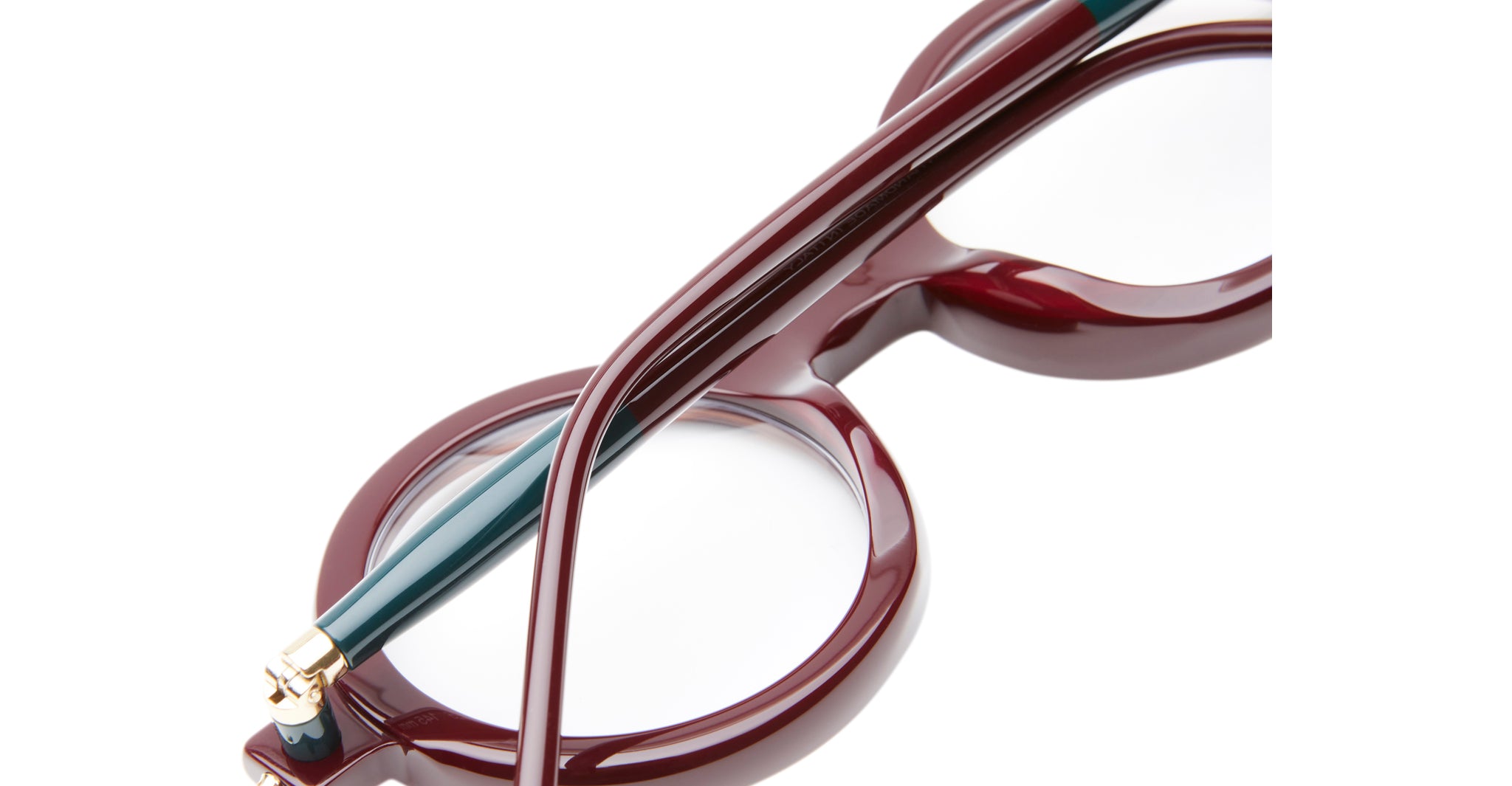 Close-up of Kuboraum P1 round eyeglasses with glossy burgundy frames and a green temple arm, set against a white background. Focus is on the frame's details and lenses.