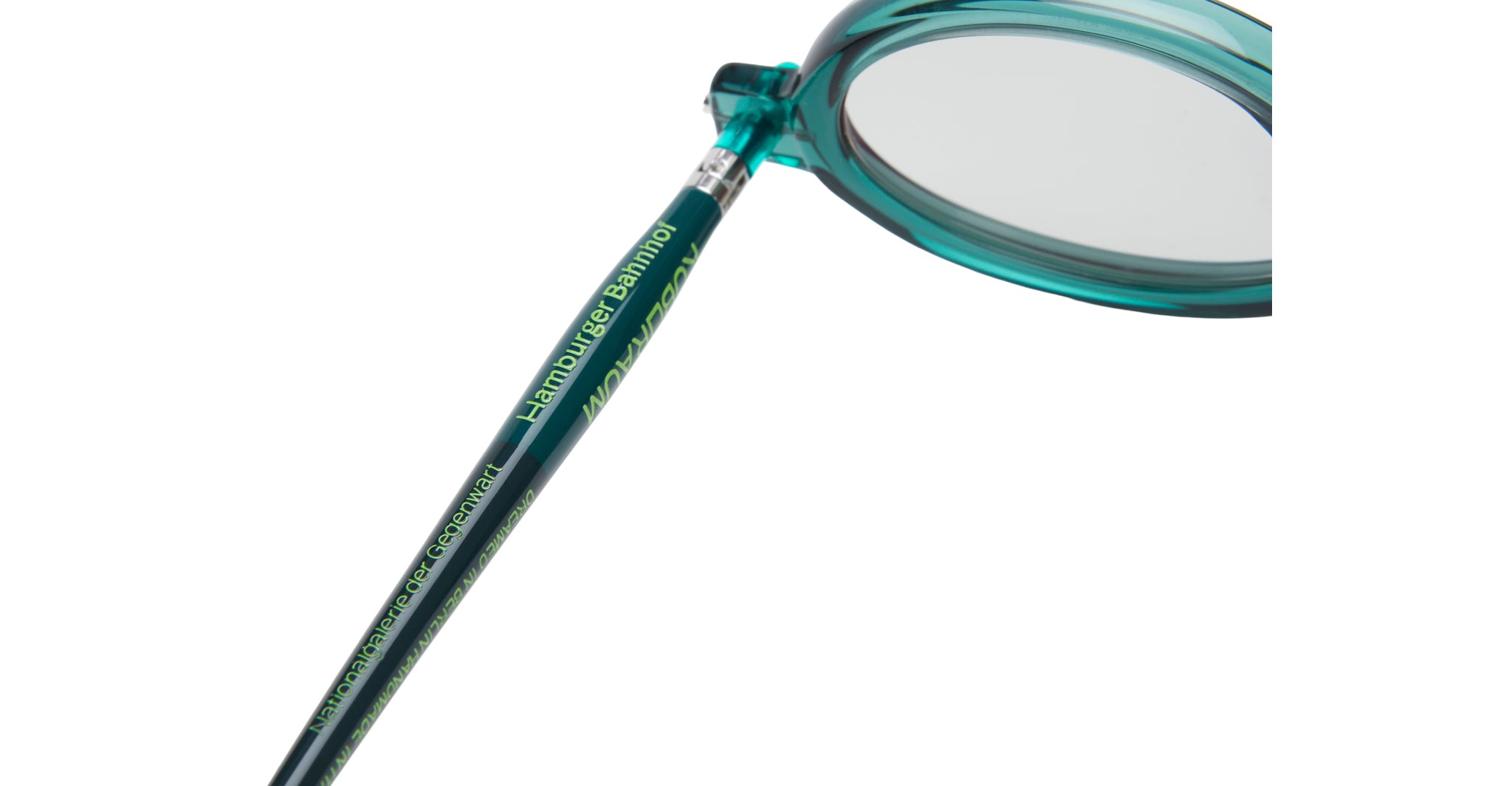 Close-up of the teal-colored arm of a pair of eyeglasses, with the words Hamburger Bahnhof and other text printed in yellow-green along the side. The clear lens and part of the round frame are visible.
