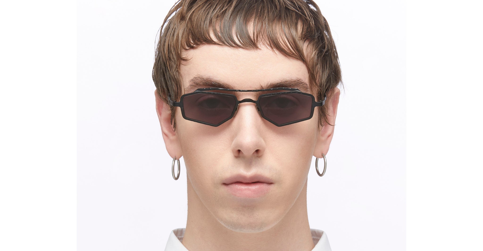 A person with short brown hair faces forward against a plain white background, wearing Kuboraum Z23 angular black sunglasses, hoop earrings, and a white shirt.