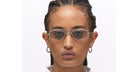 A person with braided hair wears Kuboraum Z23 geometric, silver-rimmed sunglasses and hoop earrings, gazing neutrally at the camera against a plain white background.