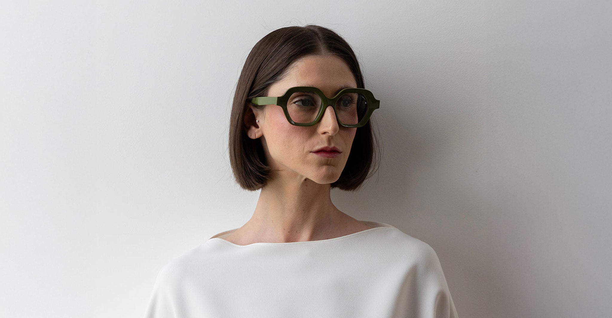 A person with a straight dark bob wears oversized green Lapima Alice glasses, standing against a plain white wall.