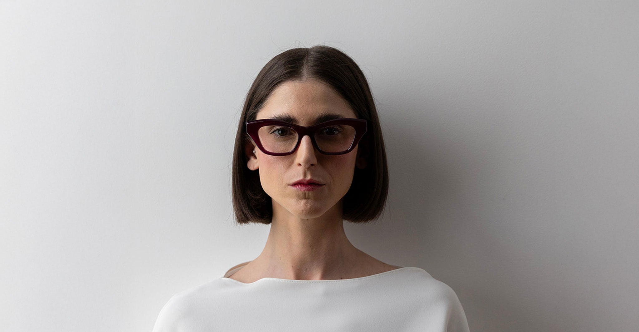 A person with straight, shoulder-length dark hair wears Lapima Bebel purple cat-eye glasses and a white top, standing against a plain white background and looking directly at the camera with a neutral expression.