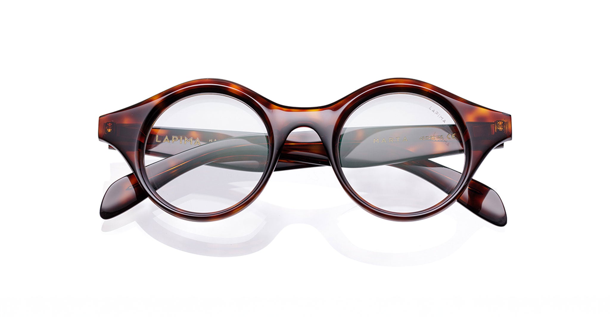 Lapima Marta eyeglasses feature a round acetate frame in tortoiseshell, handcrafted in Brazil from Italian acetate with clear lenses. The arms are crossed behind the frames, shown on a white background.