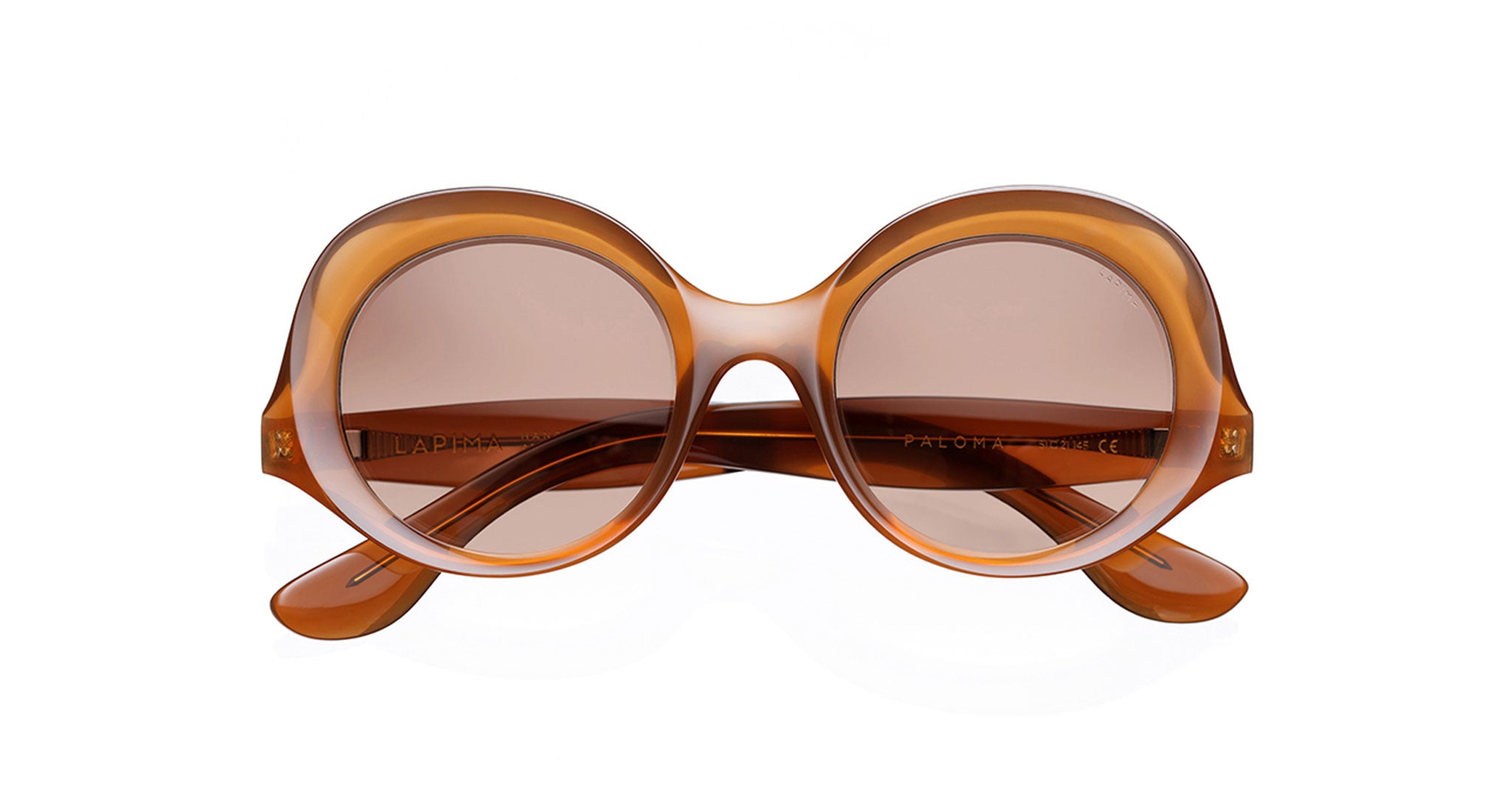 The Lapima Paloma sunglasses feature round, oversized frames in glossy amber Italian acetate with brown-tinted lenses. Handcrafted in Brazil, the arms cross stylishly behind the lenses, shown here against a white background.