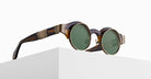 The Matsuda 10605H round sunglasses, featuring dark green lenses, brown tortoiseshell frames, and gold-toned accents on the arms, are displayed on a white minimalist pedestal against a plain background.