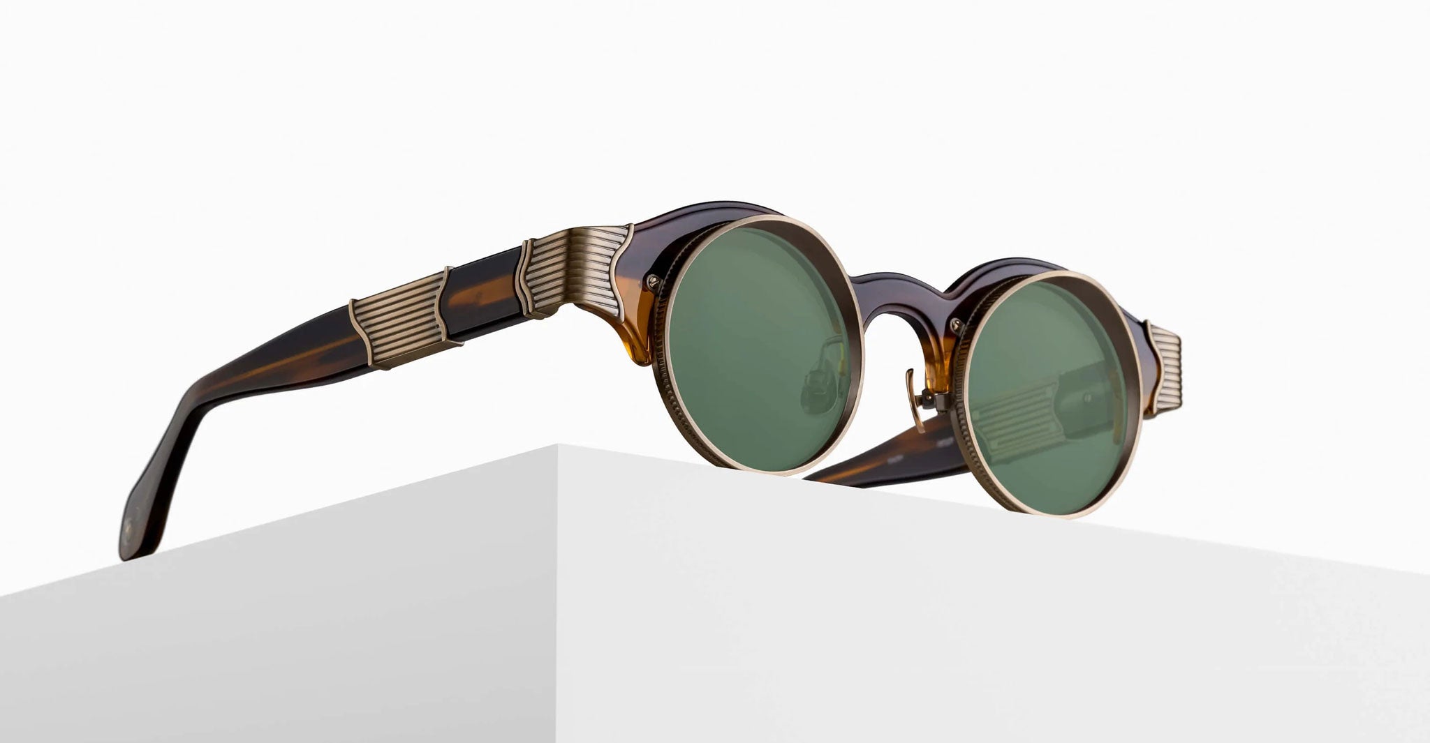 The Matsuda 10605H round sunglasses, featuring dark green lenses, brown tortoiseshell frames, and gold-toned accents on the arms, are displayed on a white minimalist pedestal against a plain background.