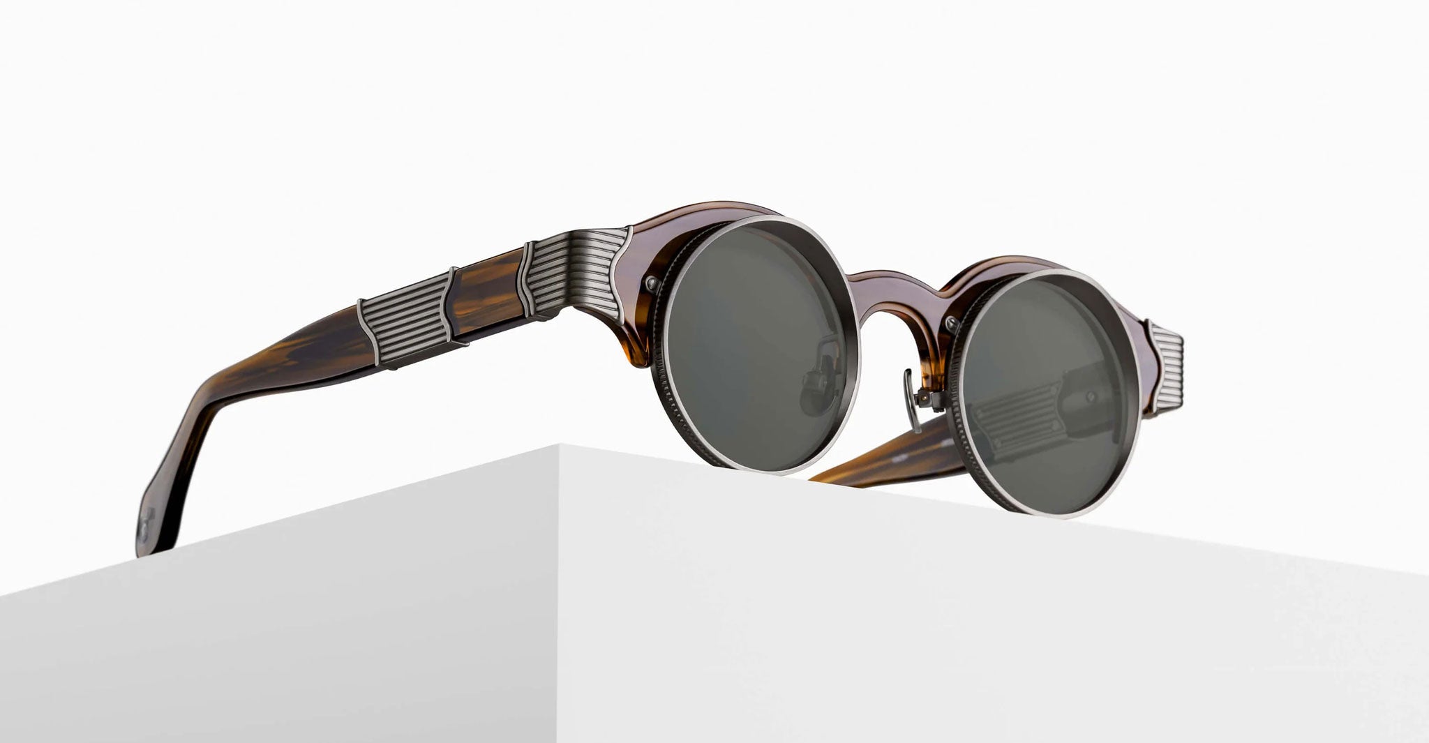 The Matsuda 10605H sunglasses feature round dark lenses and brown tortoiseshell frames, resting on a white surface. Silver metal ridged accents near the hinges on the arms add a modern, stylish touch to the design.