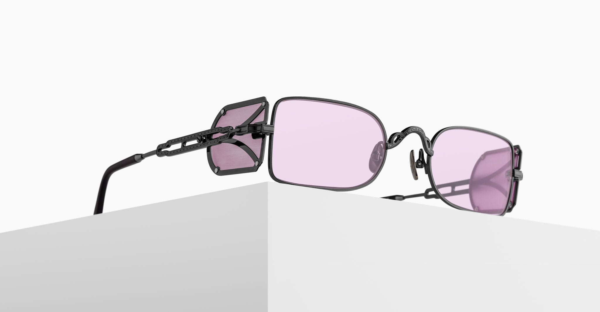 A pair of black metal sunglasses with rectangular, light purple-tinted lenses rest on the edge of a white surface, with unique side shields attached to the arms.
