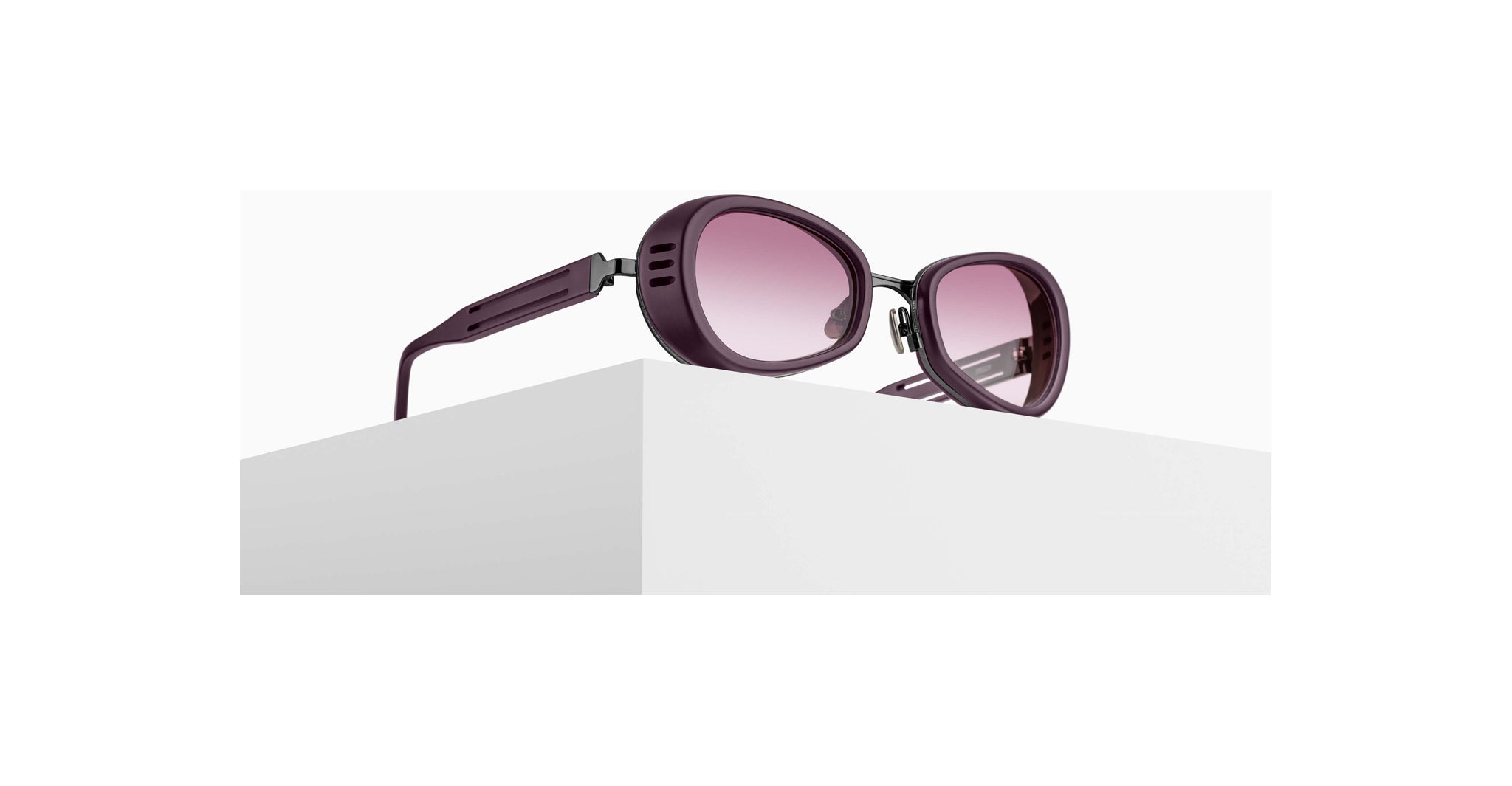 A pair of oval, purple-framed sunglasses with pink-tinted lenses is displayed on a white geometric surface against a plain white background.