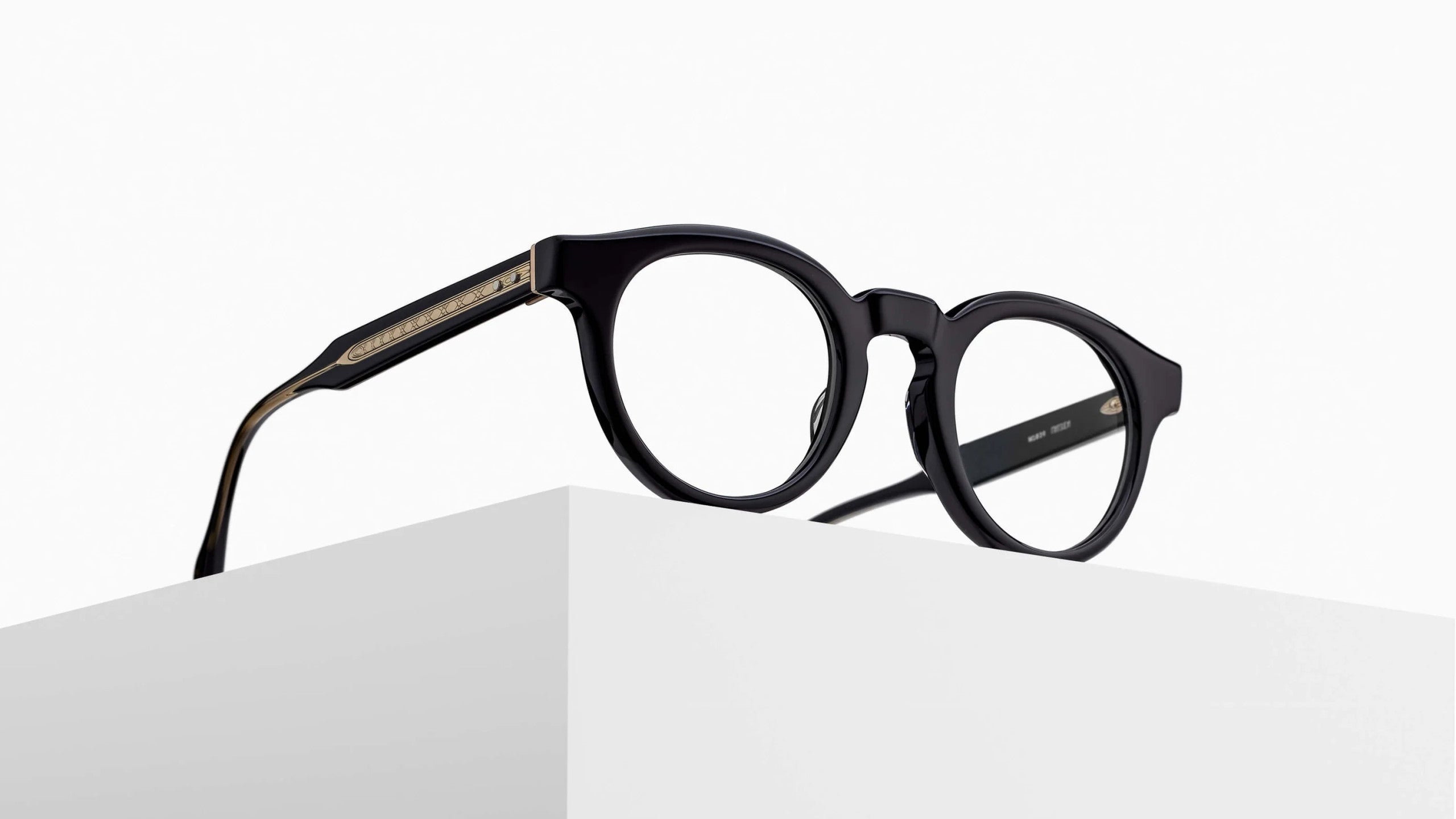The Matsuda M1036 (Copy), round black eyeglasses with thick frames, rests on the edge of a white minimalist surface against a plain background.
