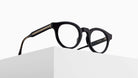 The Matsuda M1036 (Copy), round black eyeglasses with thick frames, rests on the edge of a white minimalist surface against a plain background.