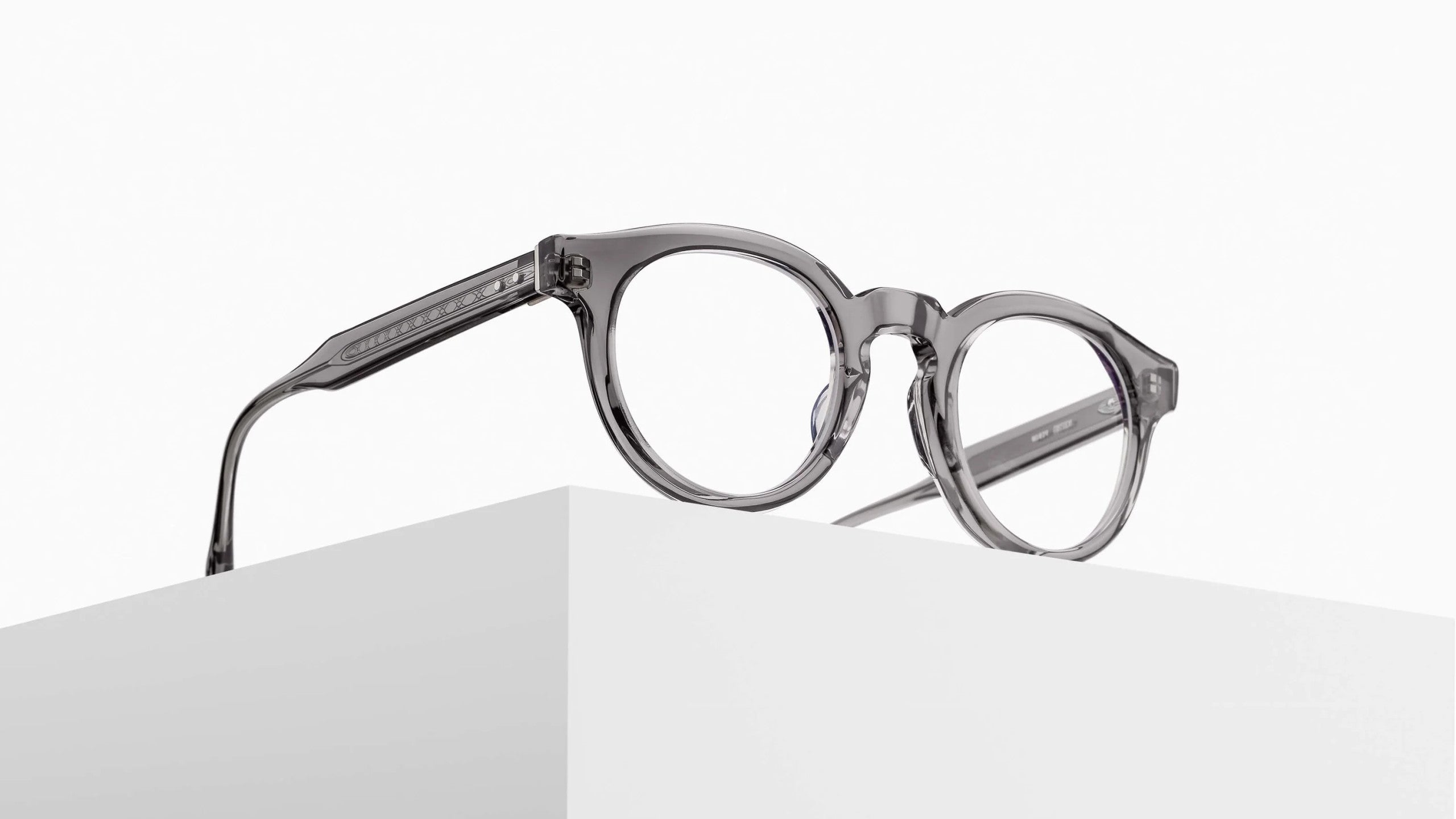 The Matsuda M1036 (Copy) round eyeglasses feature thick, transparent gray frames and are displayed atop a white geometric block against a minimalist white background.