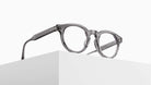 The Matsuda M1036 (Copy) round eyeglasses feature thick, transparent gray frames and are displayed atop a white geometric block against a minimalist white background.