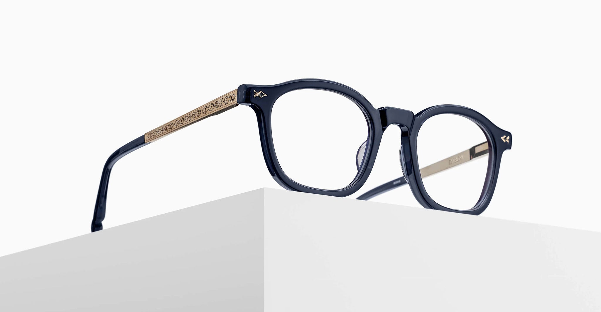 The Matsuda M2068 eyeglasses, featuring dark frames and gold, patterned arms, rest on the edge of a white minimalist surface against a plain, light background.