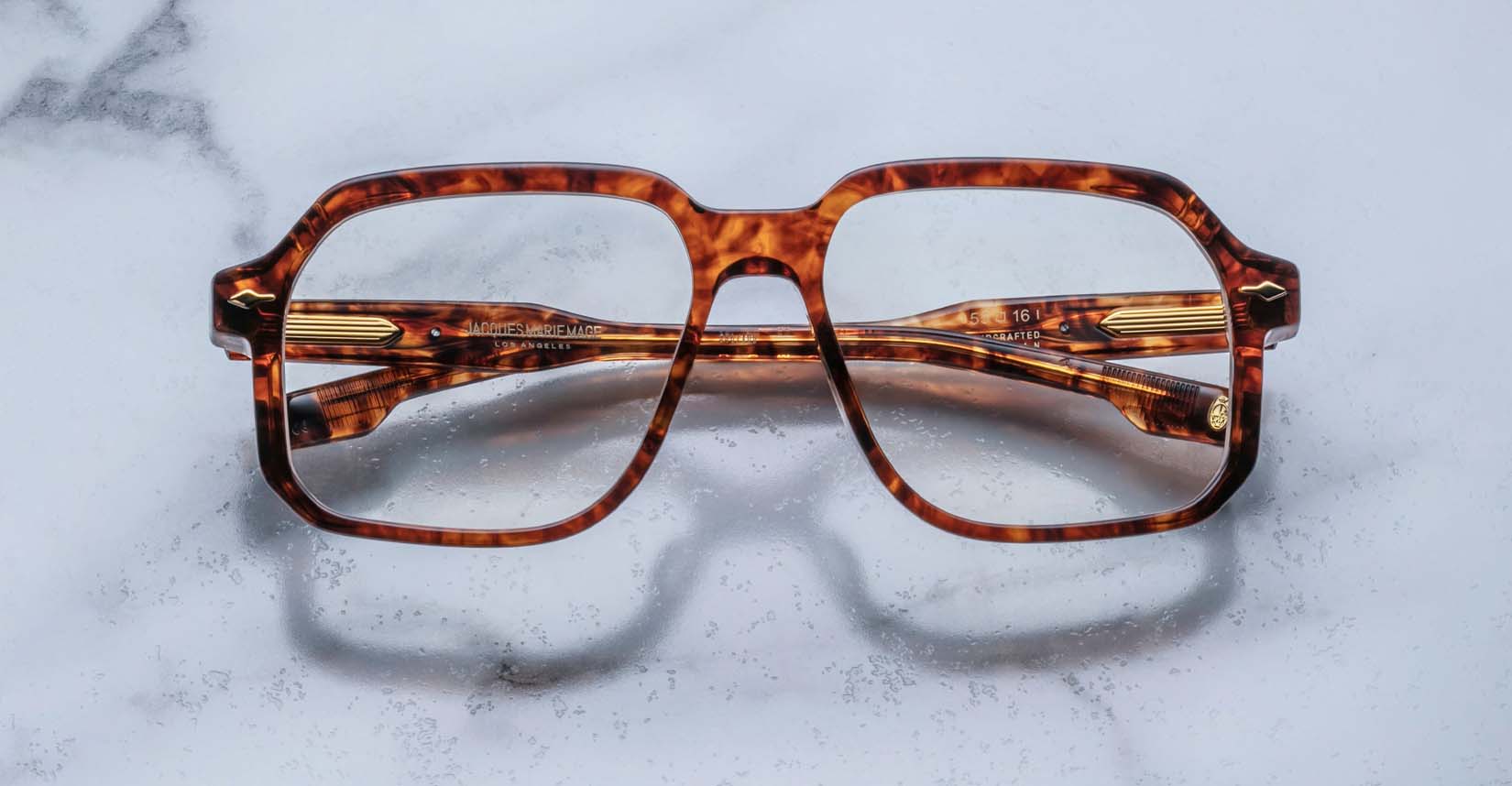 The Jacques Marie Mage Domoto eyeglasses, featuring a tortoiseshell rectangular frame and gold-detailed temples, rest on a white marble surface.