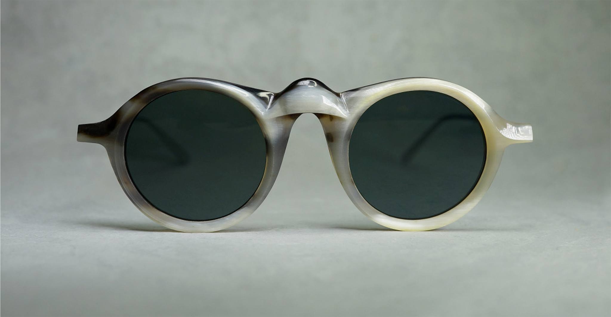 Round sunglasses with thick, ivory-colored frames and dark lenses are displayed against a light grey background. The glasses have unique, sculpted details around the nose bridge and temples.