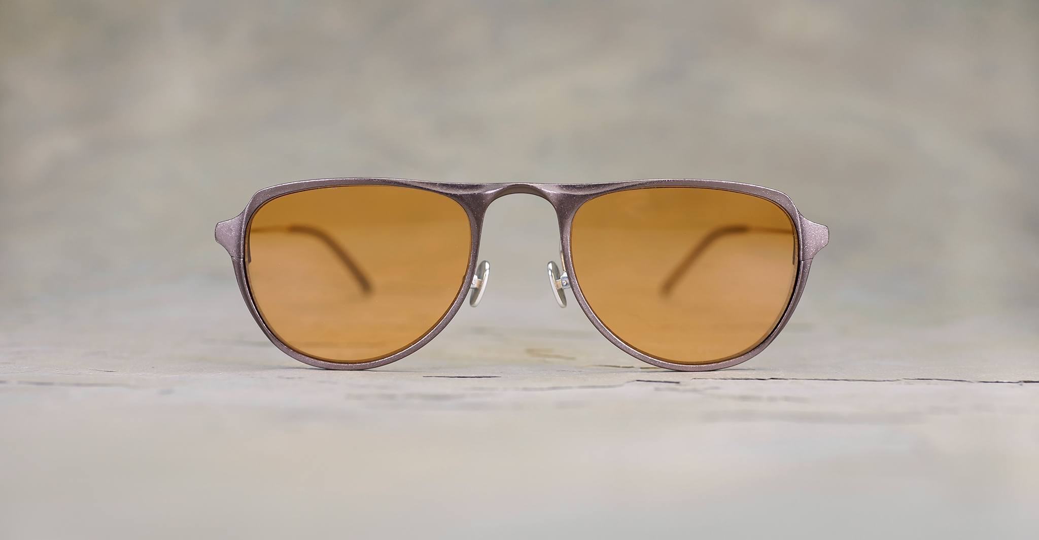 The Rigards RG0101AL aviator sunglasses, featuring amber-tinted lenses and sleek aluminum magnesium alloy frames, stand upright on a neutral surface with their arms slightly visible in the background.