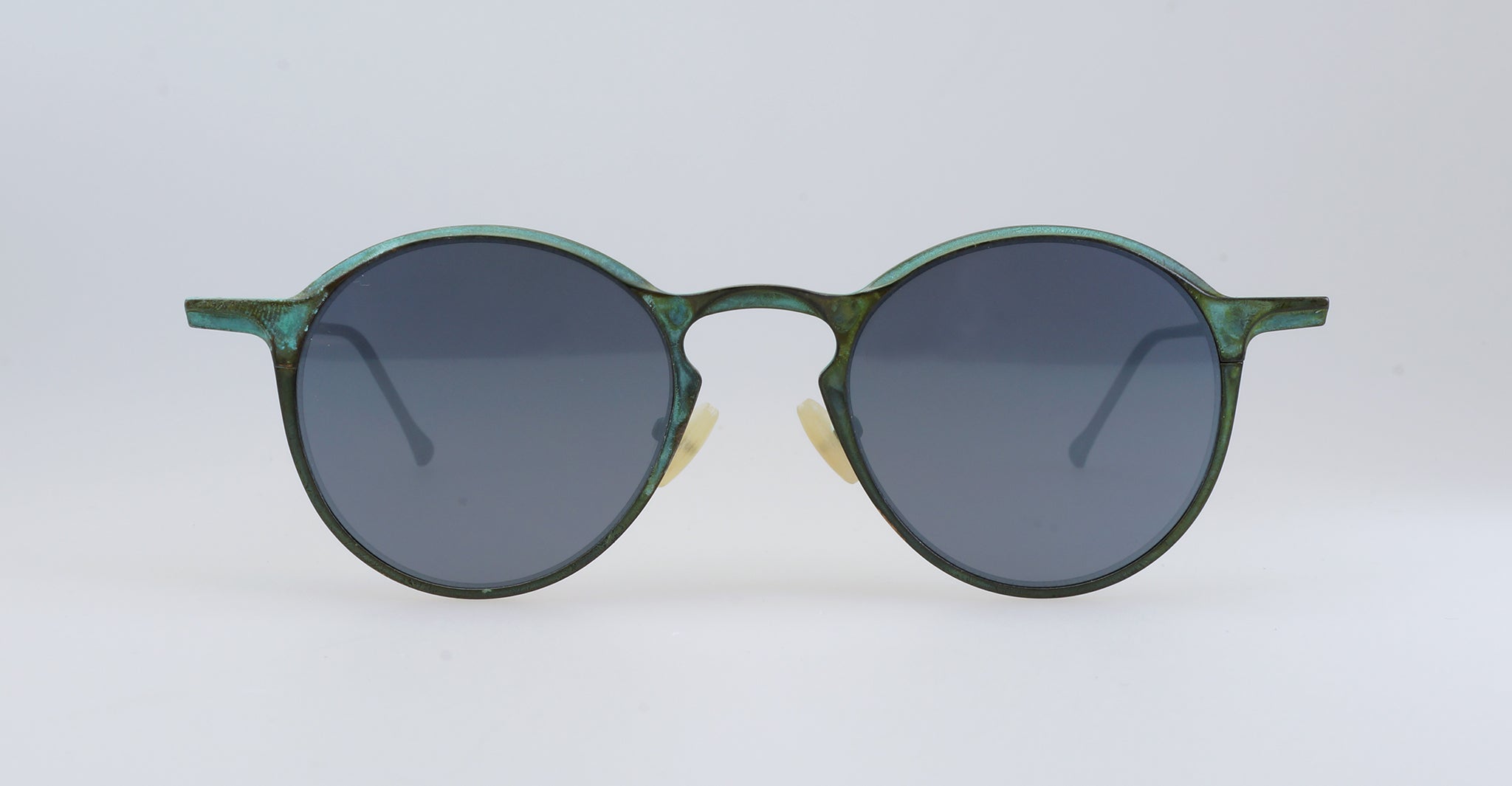 Round sunglasses with dark lenses and a thin, greenish metal frame. The temples curve outward and the nose pads are visible against a plain, light background.