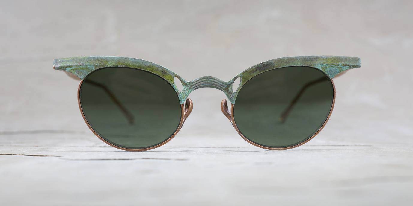A pair of vintage-style sunglasses with round dark lenses and a thick, weathered greenish frame, displayed against a light, neutral background.
