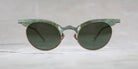 A pair of vintage-style sunglasses with round dark lenses and a thick, weathered greenish frame, displayed against a light, neutral background.