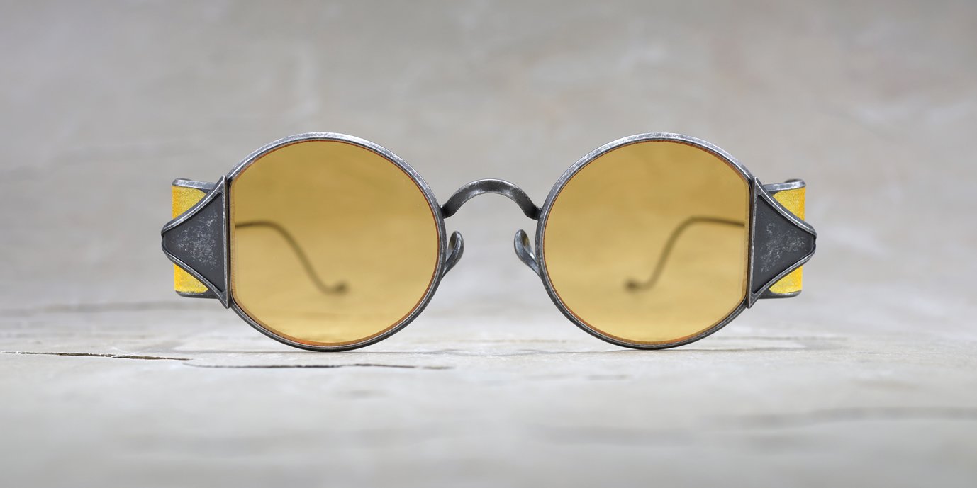 Round, vintage-style sunglasses with yellow-tinted lenses and thick, metal frames, shown from the front, placed on a light, textured surface with a blurred background.