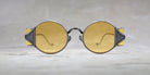 Round, vintage-style sunglasses with yellow-tinted lenses and thick, metal frames, shown from the front, placed on a light, textured surface with a blurred background.