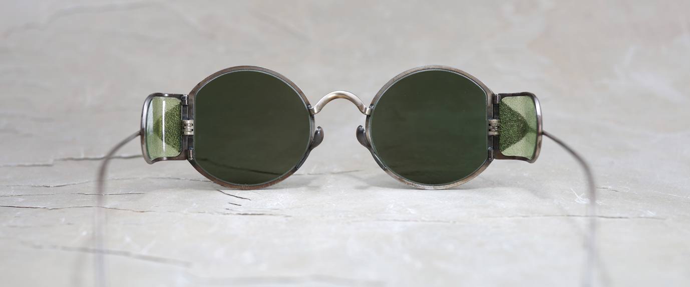 Round, vintage-style sunglasses with green-tinted lenses and side shields rest on a light, textured surface. Thin wire arms extend outward from the frames.