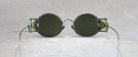 Round, vintage-style sunglasses with green-tinted lenses and side shields rest on a light, textured surface. Thin wire arms extend outward from the frames.