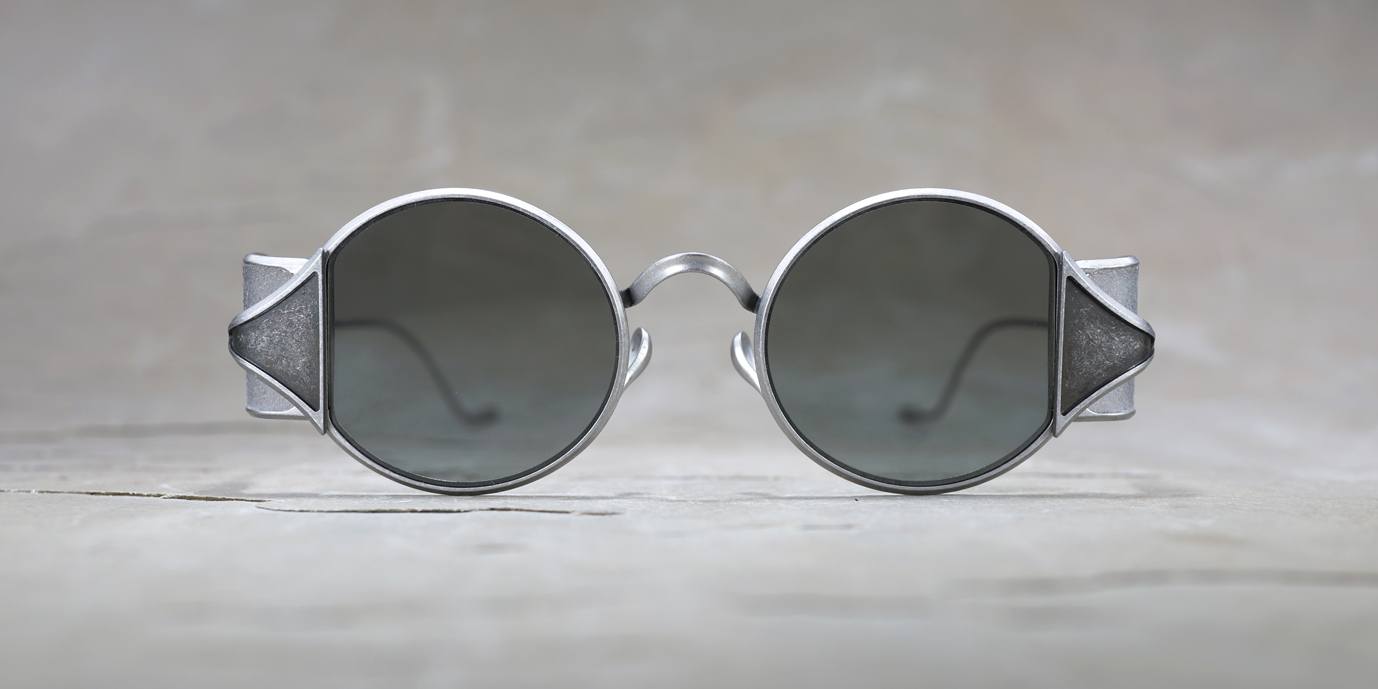 A pair of round, black-lensed sunglasses with thick, metallic silver frames, shown on a light, textured background. The sunglasses are positioned facing forward.