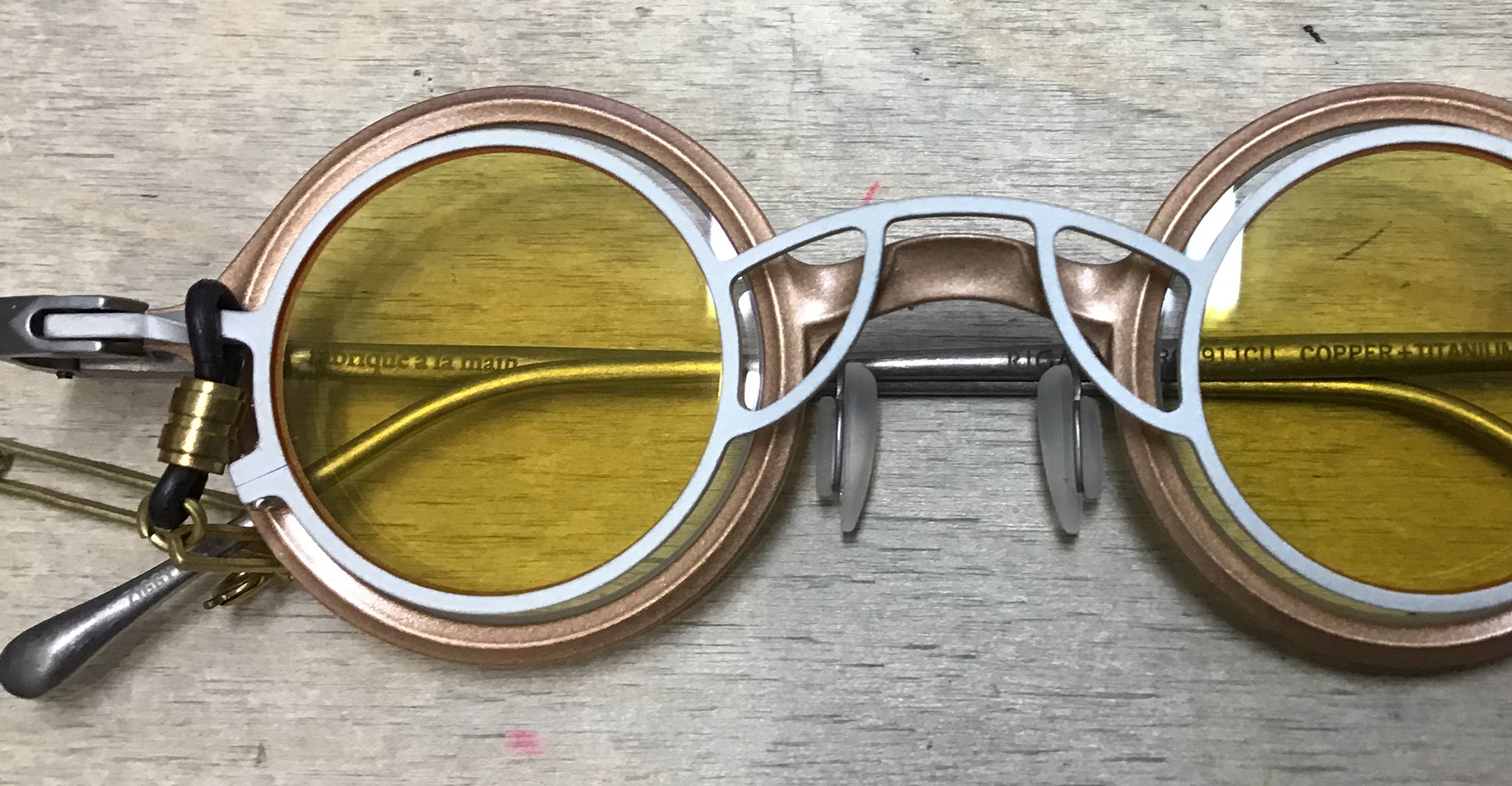A pair of round, gold and white glasses with yellow-tinted lenses sits on a light wooden surface; a black and gold clip and cord are attached to the left temple.