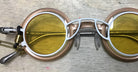 A pair of round, gold and white glasses with yellow-tinted lenses sits on a light wooden surface; a black and gold clip and cord are attached to the left temple.