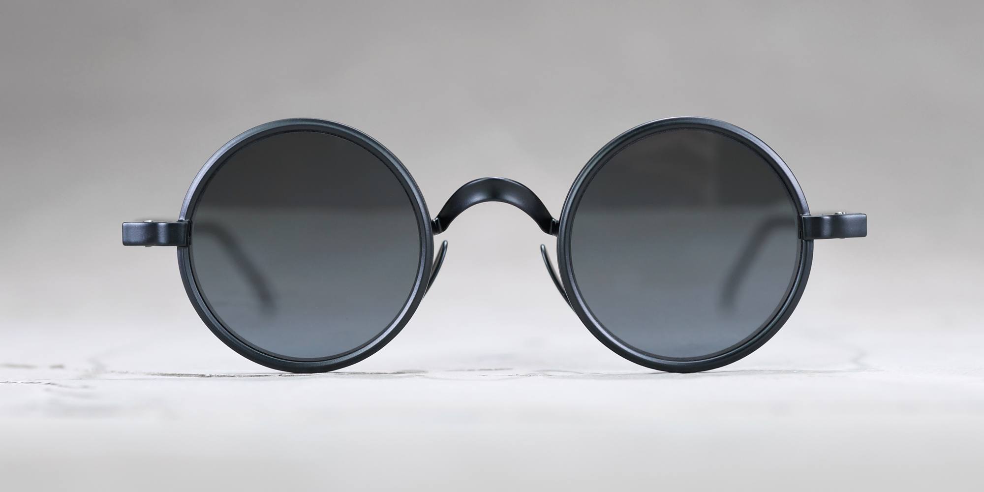 A pair of round, black sunglasses with dark tinted lenses is placed on a light, slightly textured surface, photographed from the front against a plain, neutral background.