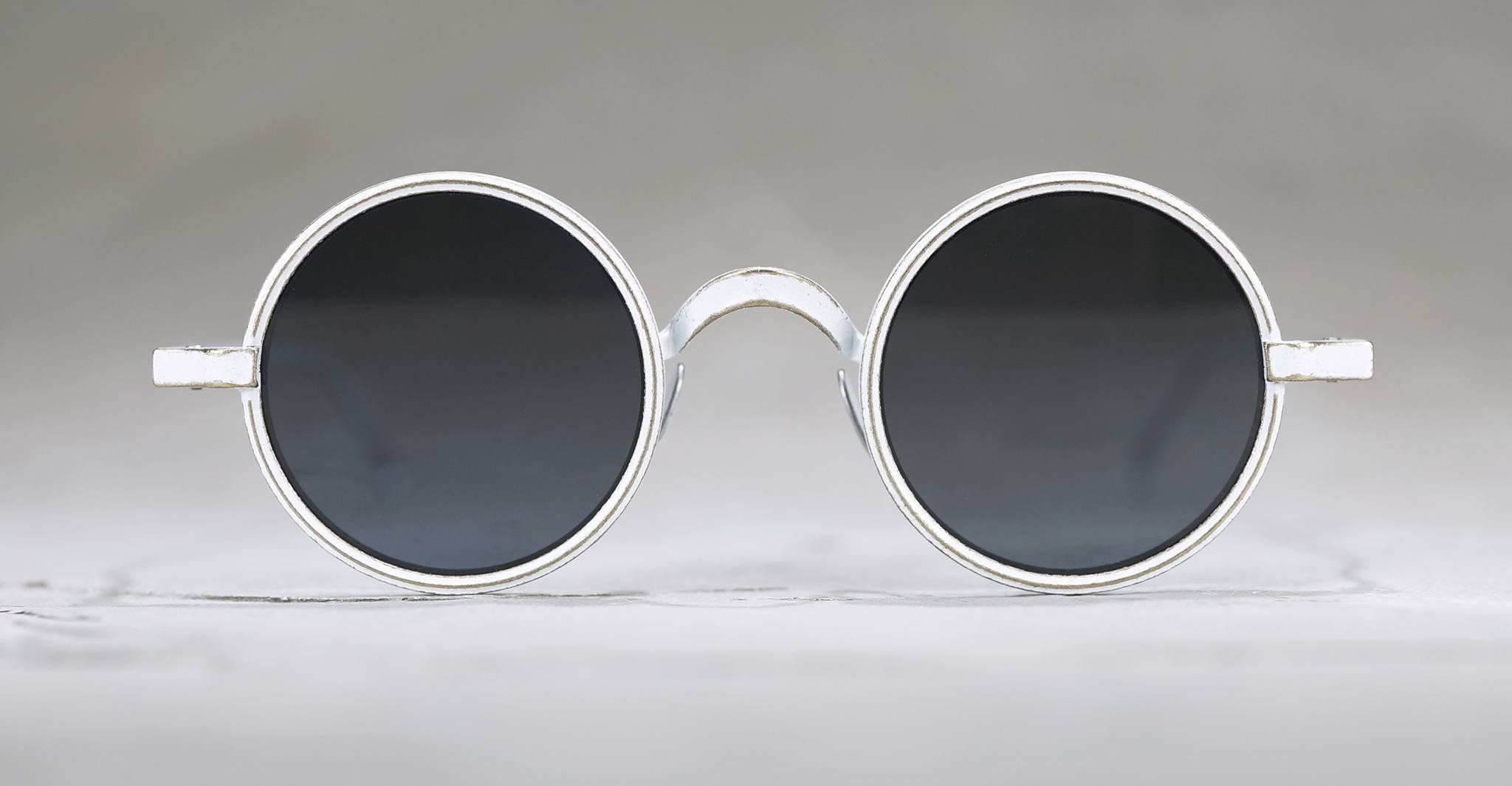 Round, black-lensed sunglasses with silver frames are centered on a light gray background, viewed from the front. The design is minimalist and retro-styled.