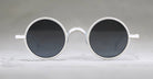 Round, black-lensed sunglasses with silver frames are centered on a light gray background, viewed from the front. The design is minimalist and retro-styled.