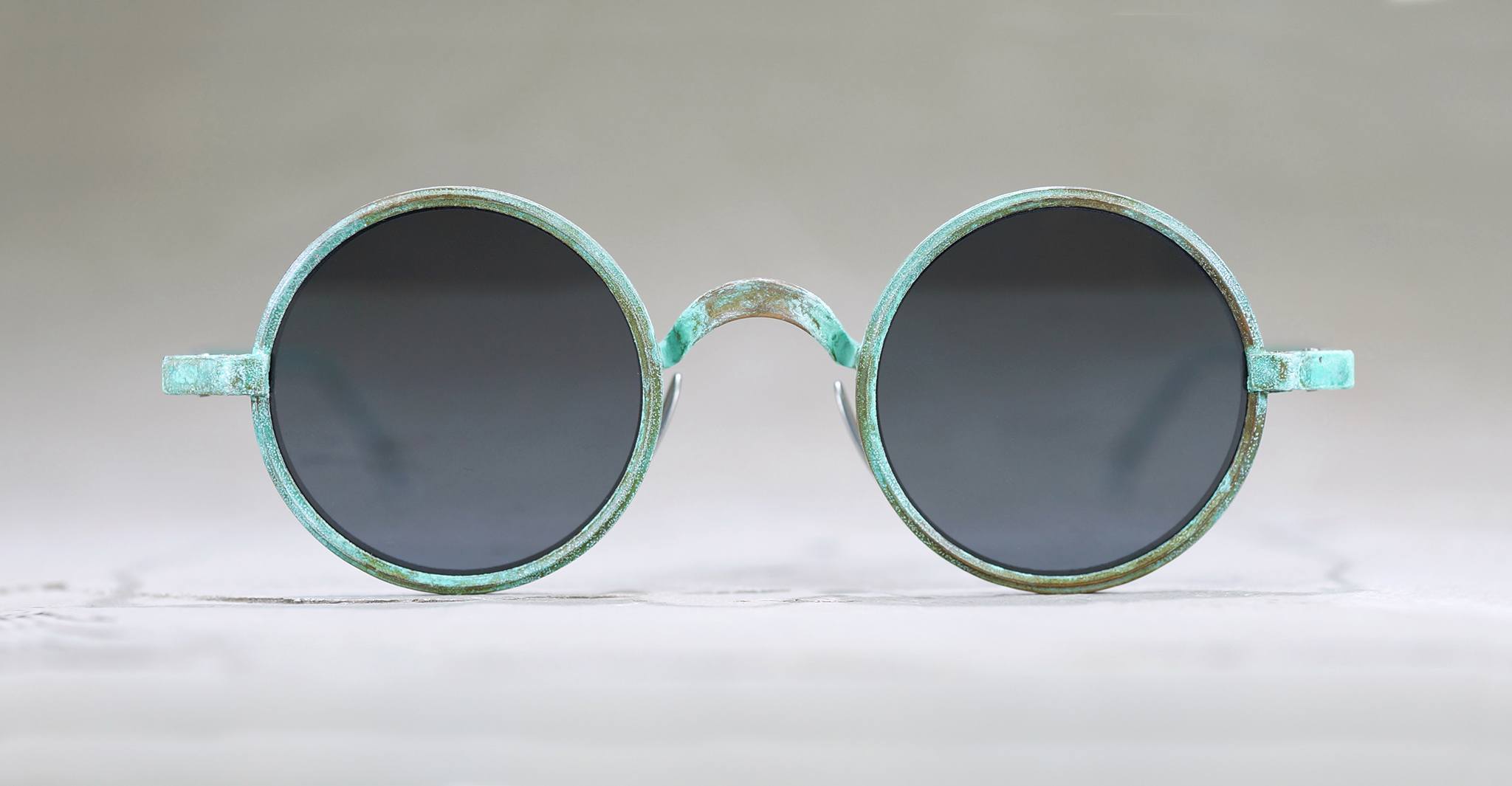Round sunglasses with dark tinted lenses and a weathered, greenish metal frame, displayed against a plain, light gray background.