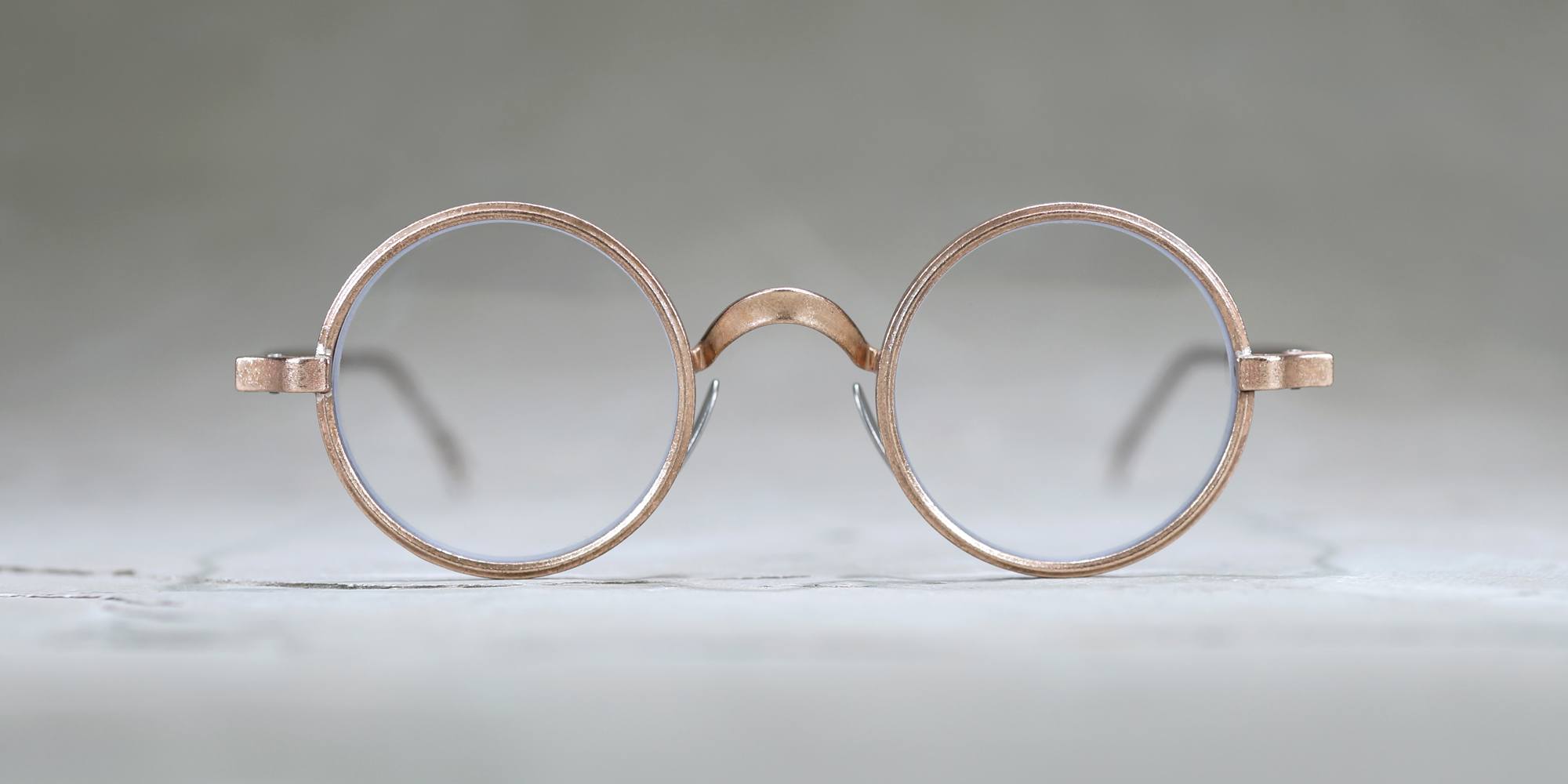 A pair of round, vintage-style eyeglasses with thin, light-colored metal frames is shown against a blurred, neutral background.