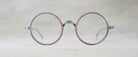 Round, vintage eyeglasses with thin, worn metal frames placed on a light, neutral background. The glasses have a slightly weathered, reddish-blue patina.