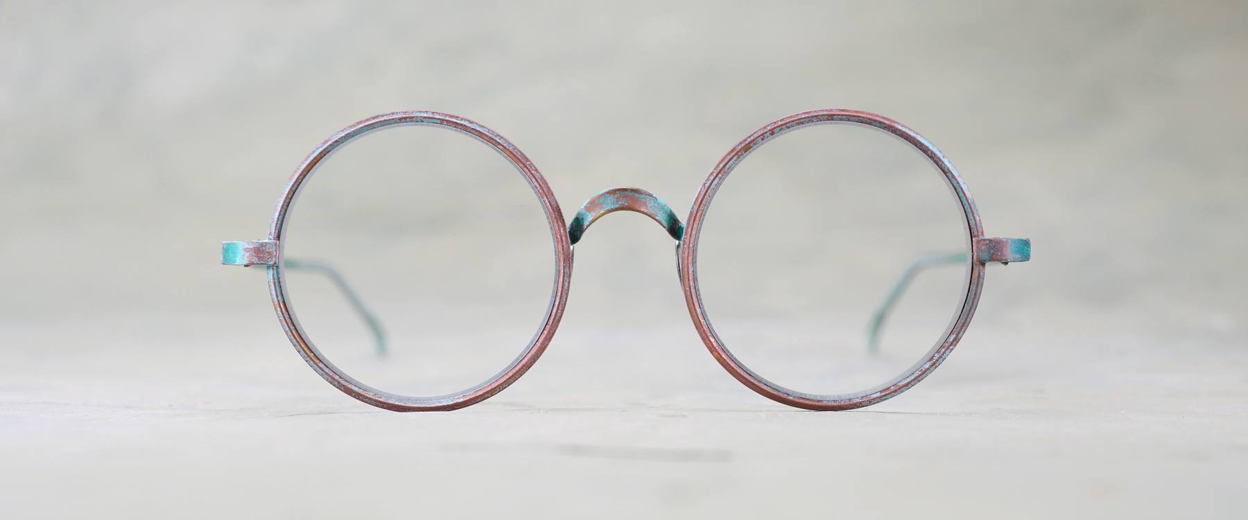 Round, vintage eyeglasses with thin, worn metal frames placed on a light, neutral background. The glasses have a slightly weathered, reddish-blue patina.