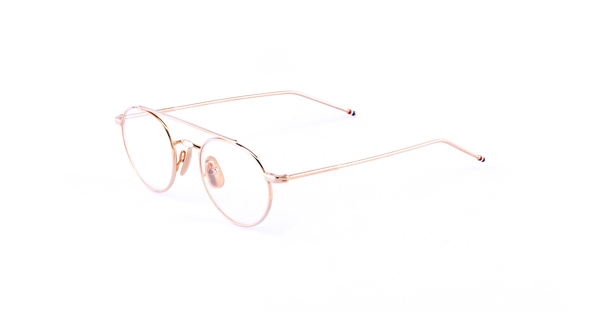 A pair of round, thin metal eyeglasses with rose gold frames and clear lenses rests on a white background. The temples have subtle colored tips, showcasing the refined style of Thom Browne 101 eyeglasses in Rose Gold.