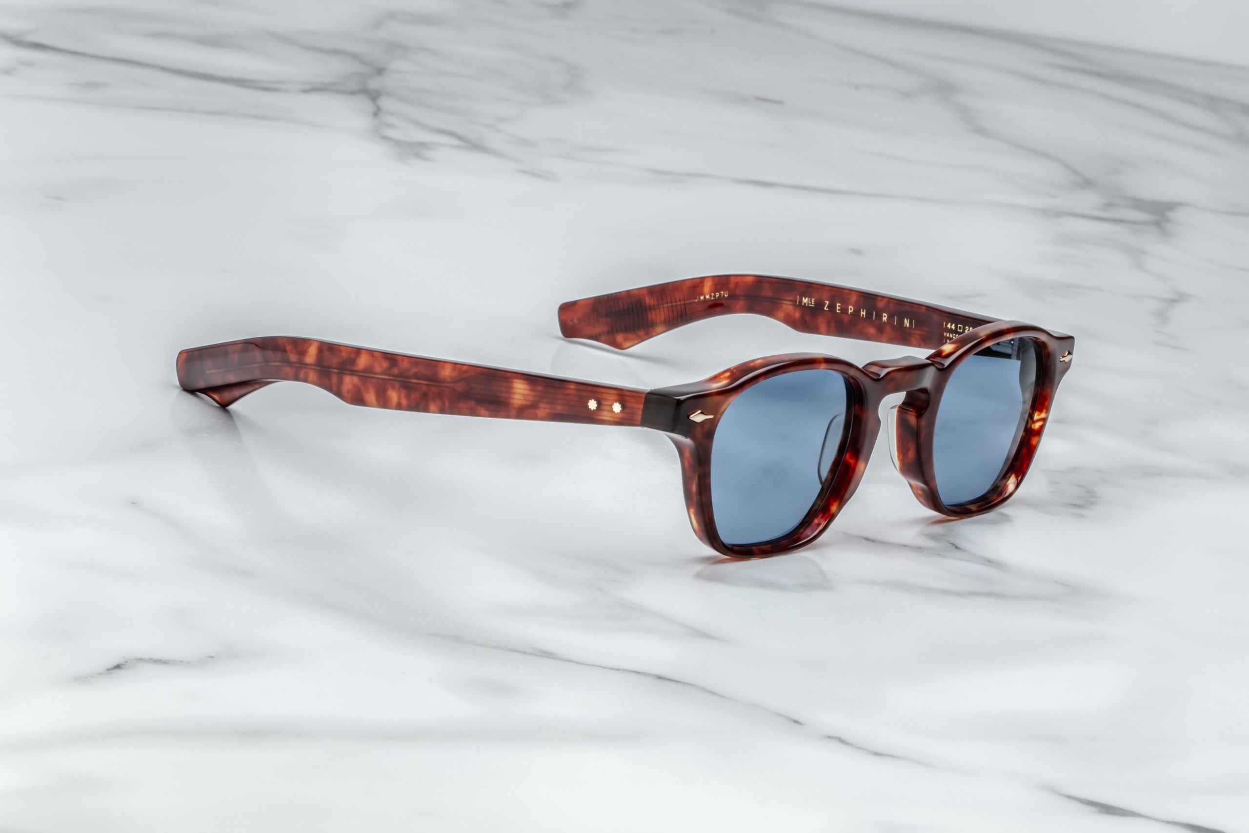 Jacques Marie Mage Zephirin limited edition acetate spectacles with blue lenses are showcased on a white marble surface—exclusive eyewear for discerning collectors.