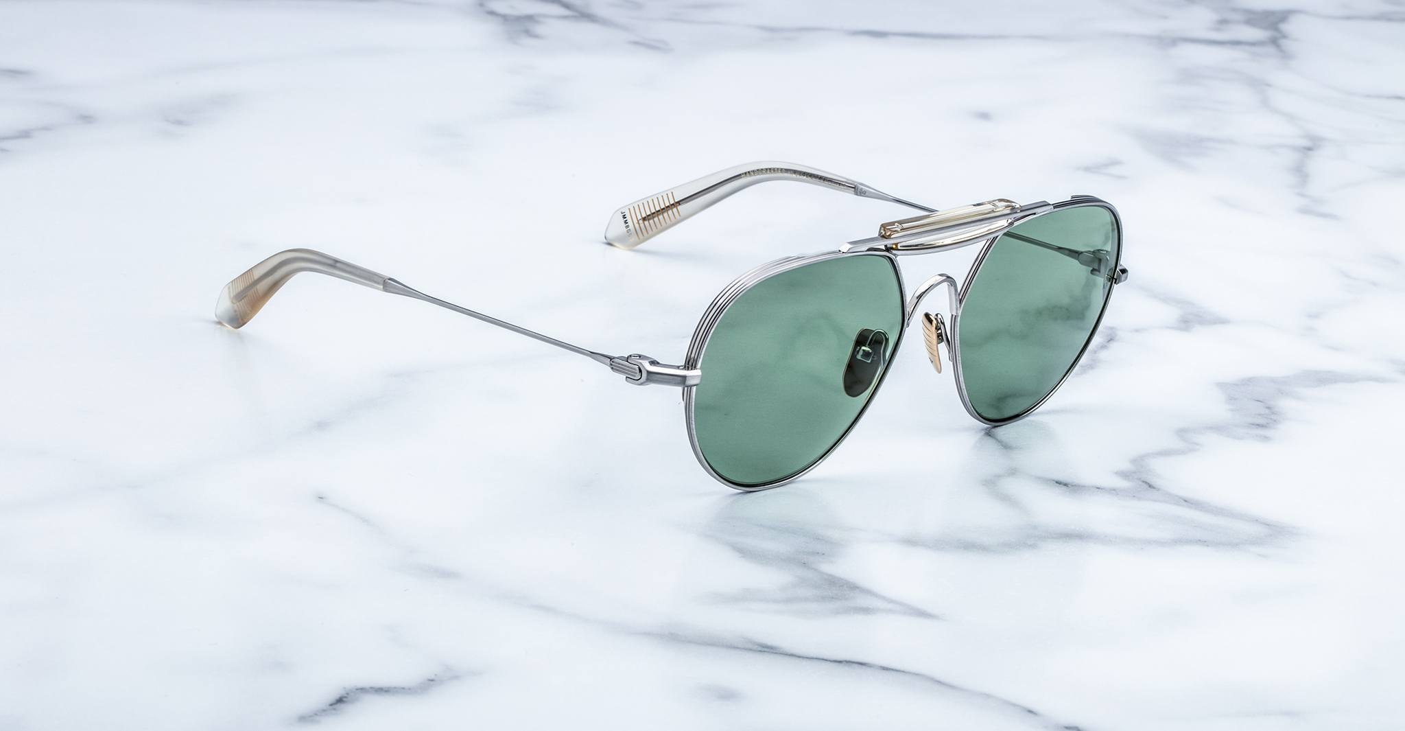 Silver-framed aviator sunglasses with green-tinted lenses resting on a white marble surface with subtle gray veining.