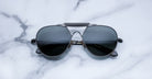 A pair of black aviator sunglasses with dark lenses lies on a white marble surface with gray veining.