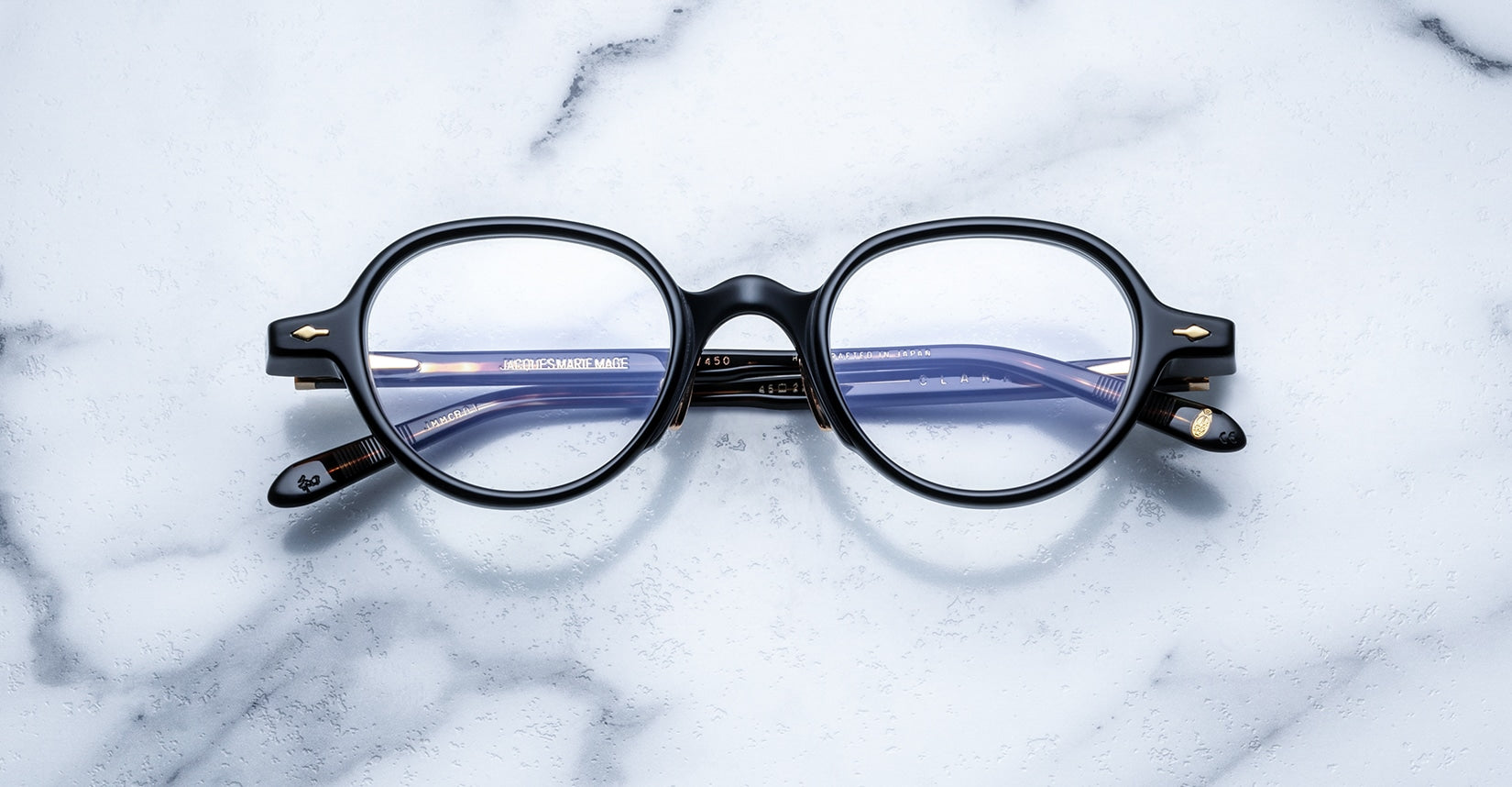The Jacques Marie Mage Clark eyeglasses by Jacques Marie Mage, featuring black round frames and blue-tinted lenses inspired by midcentury style, rest on a white marble surface.