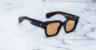 Black-framed, square-shaped sunglasses with brown-tinted lenses rest on a white marble surface. The frames have a glossy finish with slight tortoiseshell pattern visible on the inner arms.