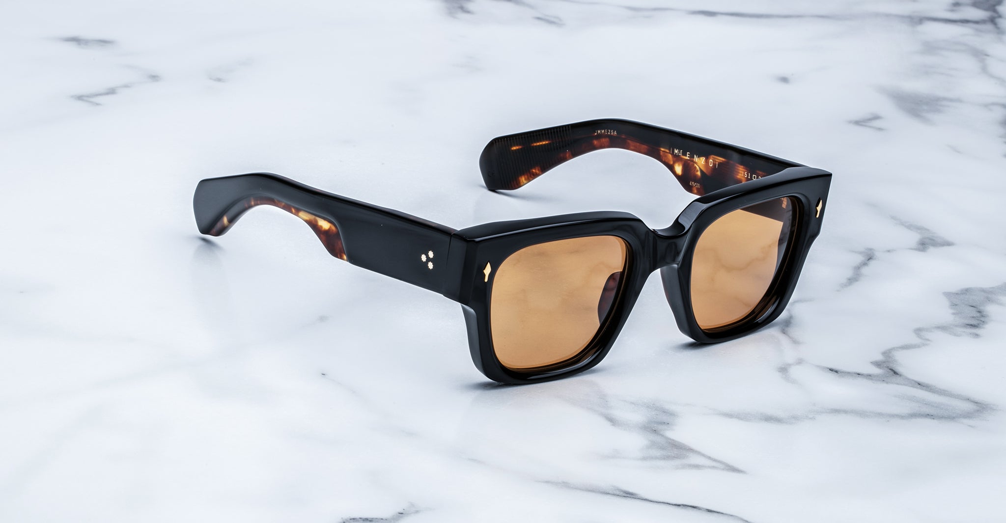 Black-framed, square-shaped sunglasses with brown-tinted lenses rest on a white marble surface. The frames have a glossy finish with slight tortoiseshell pattern visible on the inner arms.