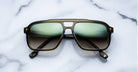 A pair of olive-green sunglasses with large, rectangular gradient lenses rests on a white marble surface with gray veining.