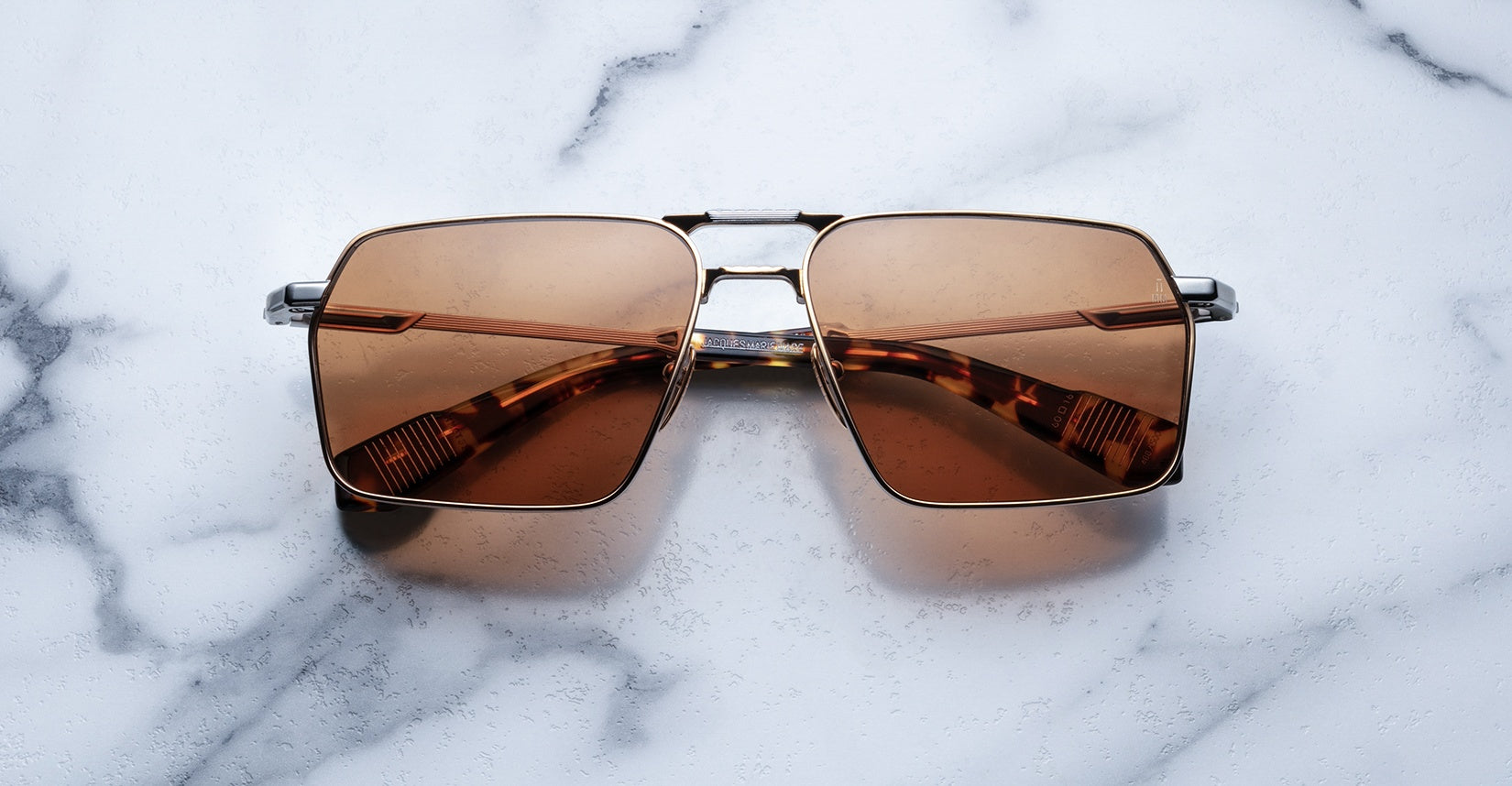 The Jacques Marie Mage GT features brown lenses and a square metallic frame, showcased on a white marble surface. These sunglasses offer a stylish, modern design.