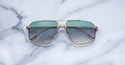 Jacques Marie Mage Jagger sunglasses by Jacques Marie Mage, featuring gold-tone oversized aviator frames with green gradient lenses, displayed elegantly on a white marble surface.