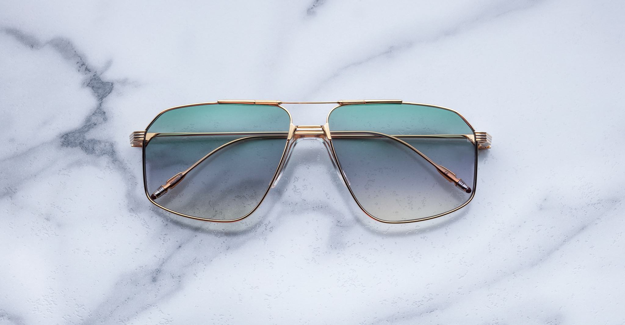 Jacques Marie Mage Jagger sunglasses by Jacques Marie Mage, featuring gold-tone oversized aviator frames with green gradient lenses, displayed elegantly on a white marble surface.