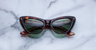 A pair of cat-eye sunglasses with dark green lenses and a brown tortoiseshell frame rests on a light gray marble surface.